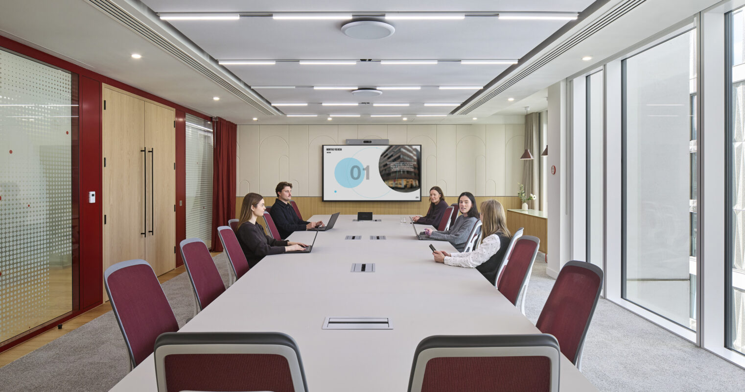 A modern conference room features a long, minimalist table surrounded by ergonomic chairs in maroon upholstery. Attendees engage with laptops and tablets, while a large screen displays a presentation. Natural light streams in through expansive glass walls, enhancing the contemporary, collaborative atmosphere. Elegant acoustic panels add visual interest while improving sound quality.