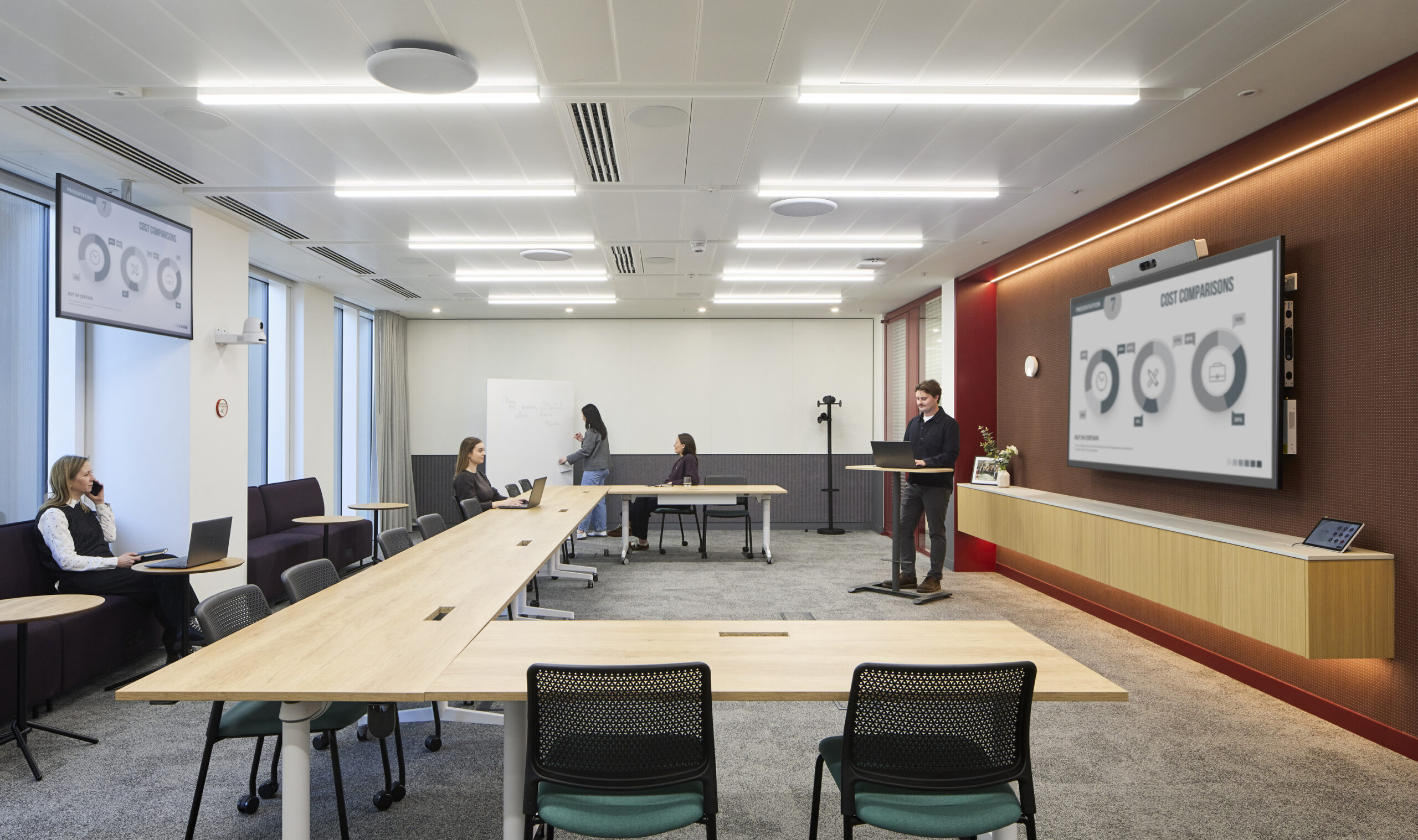 Modern conference room featuring collaborative seating arrangements and streamlined tables. A central presenter stands at a podium, with digital screens displaying information. Natural light floods the space through large windows, complemented by sound-absorbing elements and neutral-colored furnishings, fostering an inviting and efficient work environment.