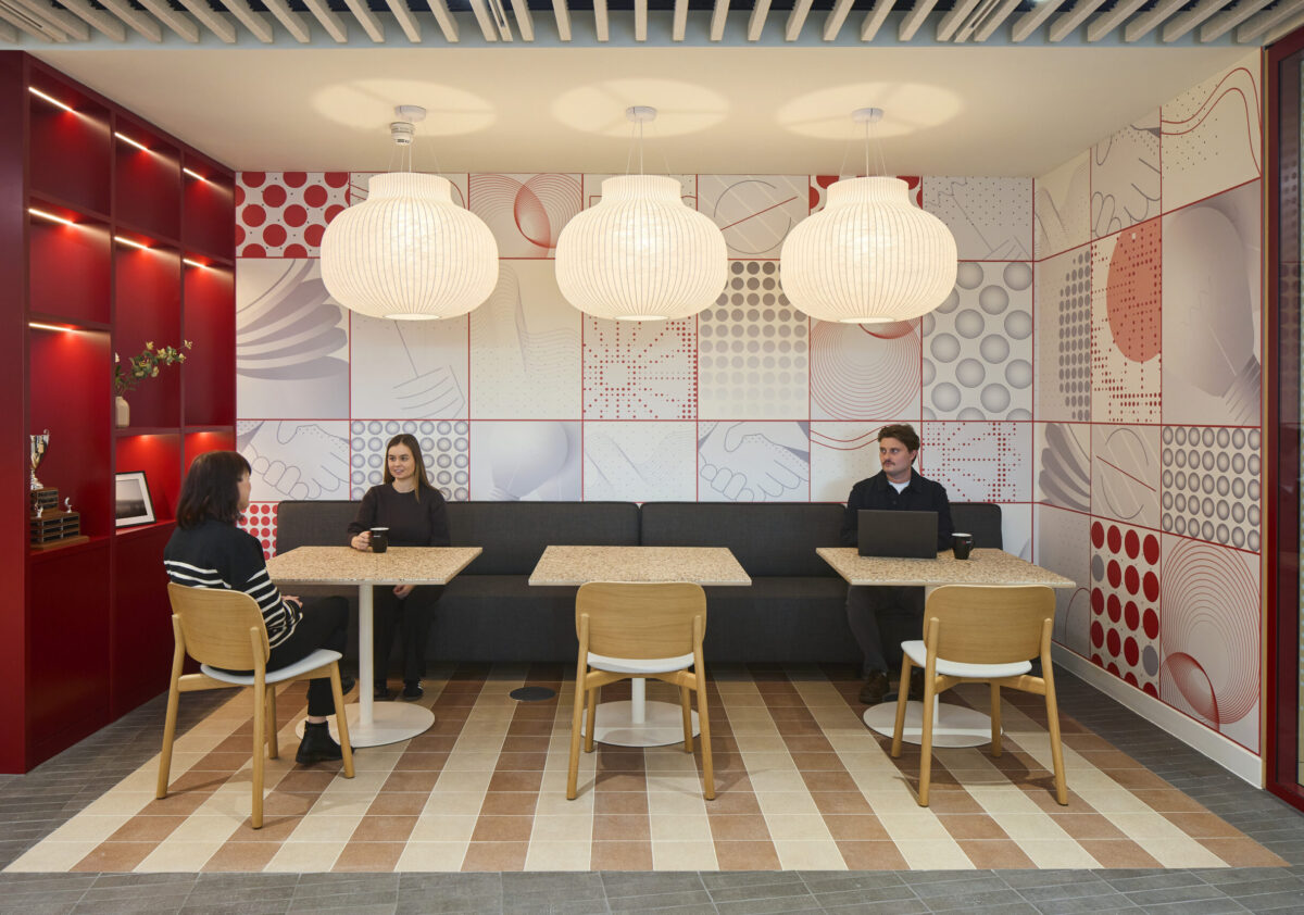 A modern collaboration space features a striking red accent wall adorned with abstract graphic patterns, complemented by elegant pendant lighting. The room includes a versatile seating arrangement: a plush gray banquette paired with light wood tables and chairs, fostering an inviting atmosphere for casual meetings and social interaction.