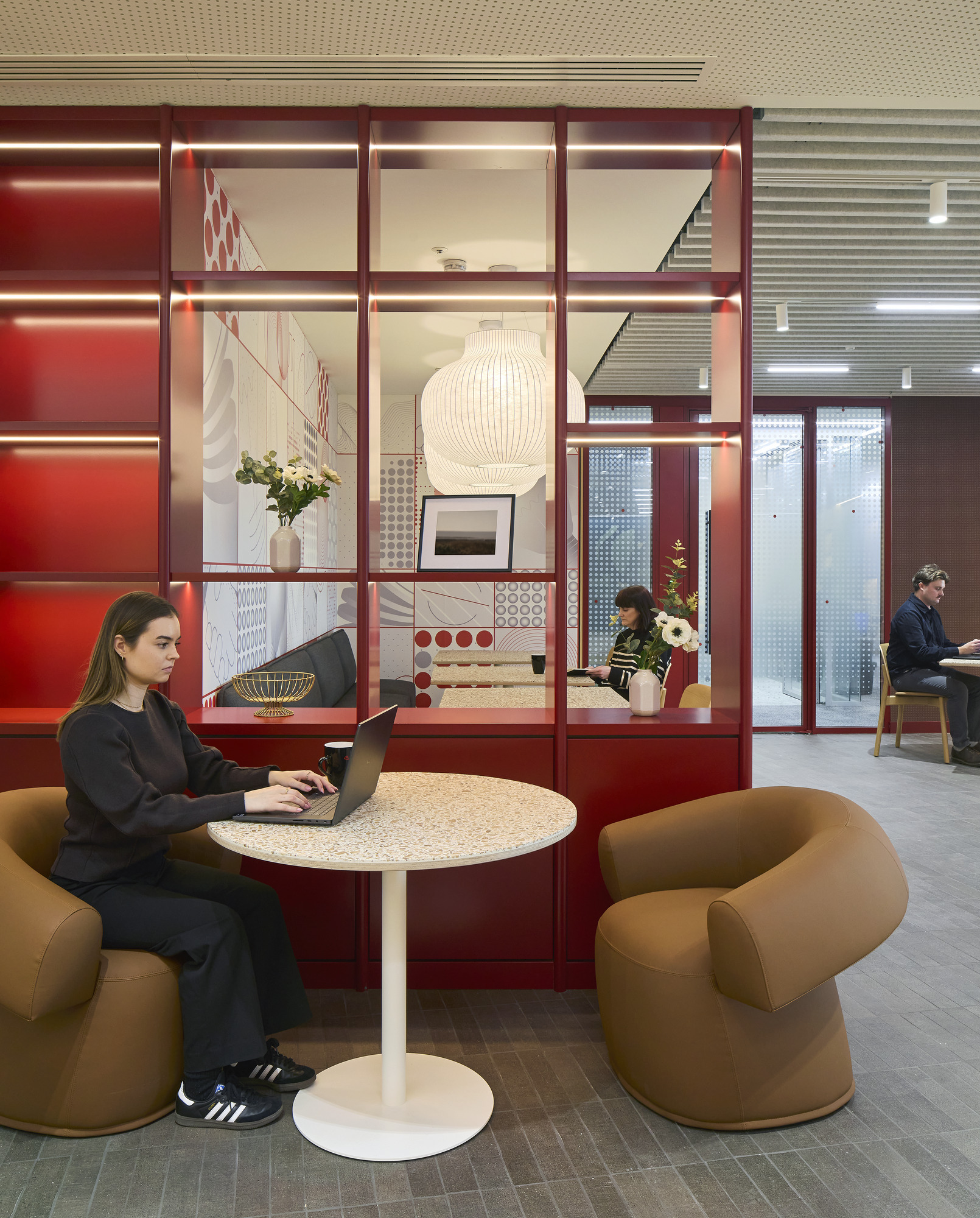 A modern workspace features a woman focused on her laptop at a round, speckled table. Plush, curved brown chairs provide comfort. Bright red partitions with integrated lighting, patterned walls, and a large pendant lamp enhance the dynamic atmosphere, promoting collaboration while ensuring privacy within the open layout.