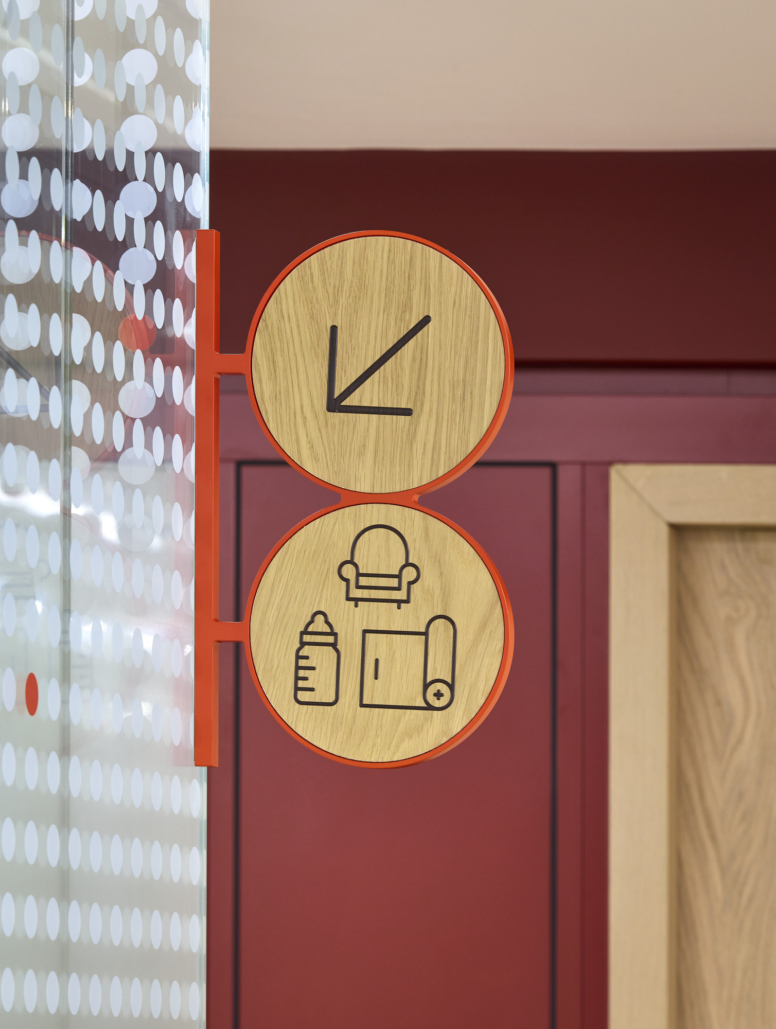Wooden directional signage prominently features minimalist icons, guiding visitors to a seating area, baby supplies, and changing facilities. The circular design, framed in vibrant orange, contrasts with the rich burgundy wall, while the playful yet professional aesthetic enhances the overall interior ambiance and user experience.