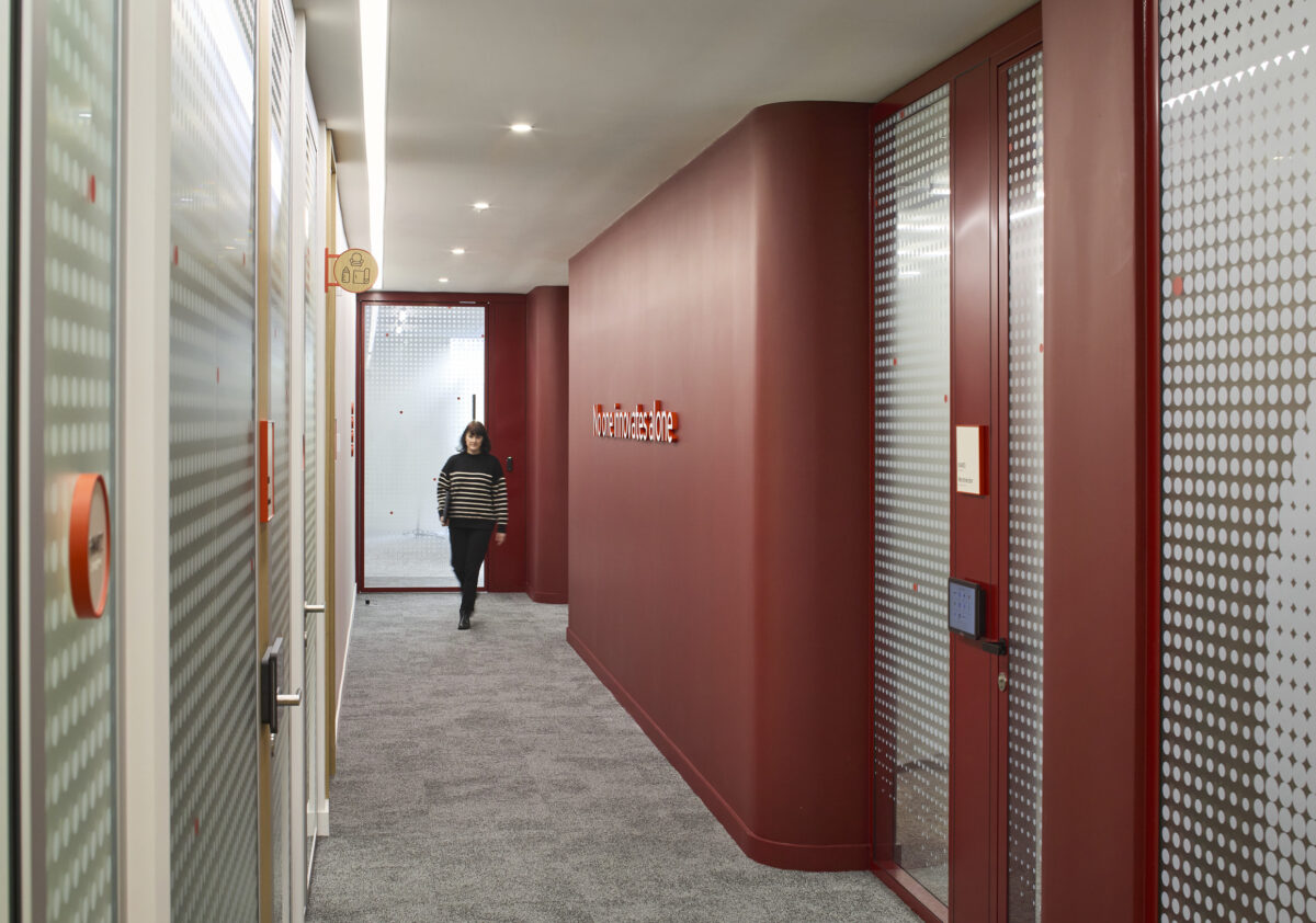 A narrow hallway features a vibrant red accent wall and modern frosted glass office partitions with dotted patterns. Soft lighting enhances the contemporary ambiance. A person walks past the glass doors, which display a motivational phrase, exemplifying a blend of functionality and aesthetic appeal in a professional setting.
