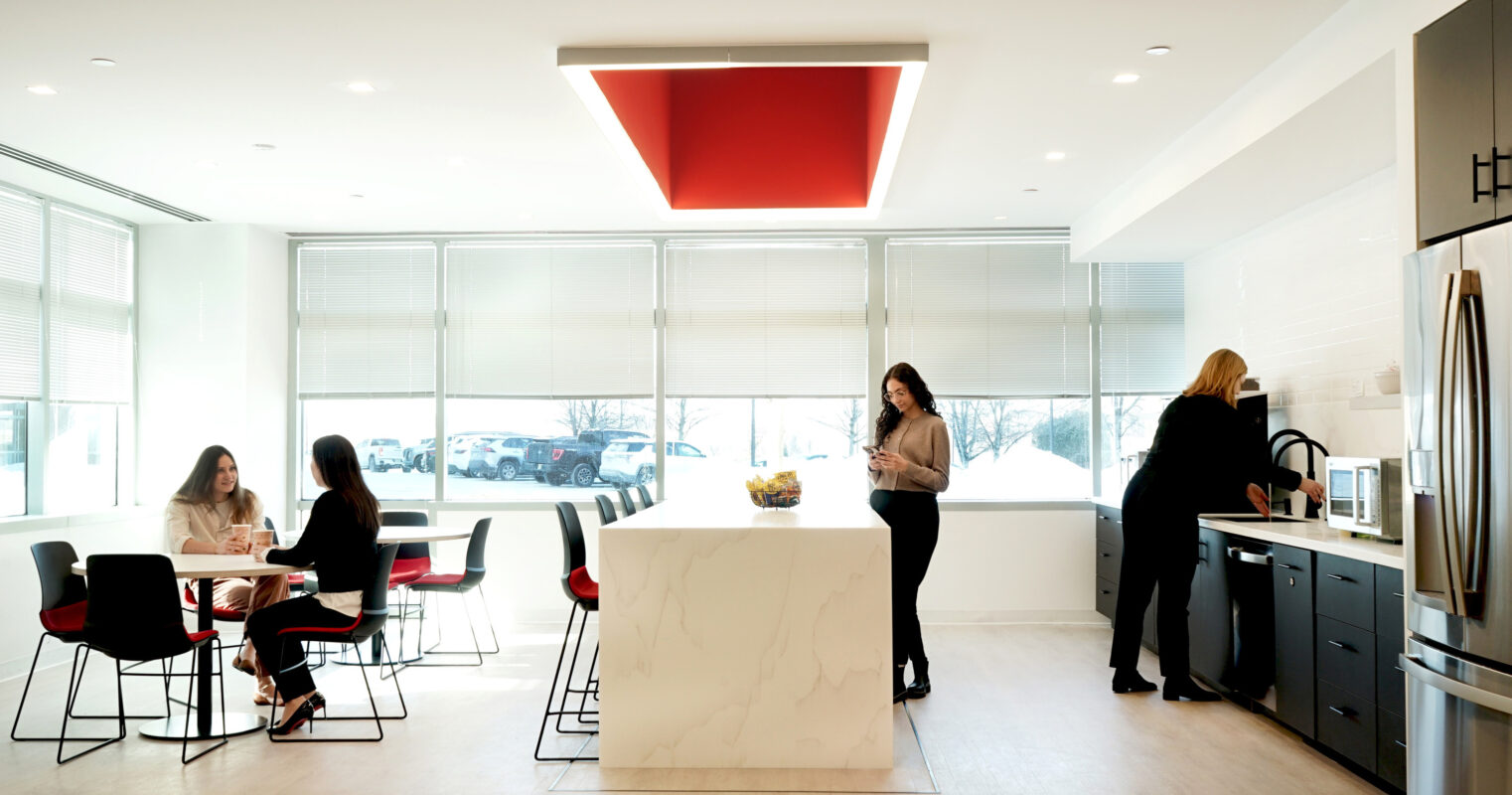 Modern office kitchen featuring a large white island with a striking red ceiling highlight. Collaborative seating area with sleek black and red chairs facilitates teamwork. Well-equipped kitchen area boasts stainless steel appliances and minimalistic cabinetry, enhancing functionality and aesthetic appeal in a bright, open environment.
