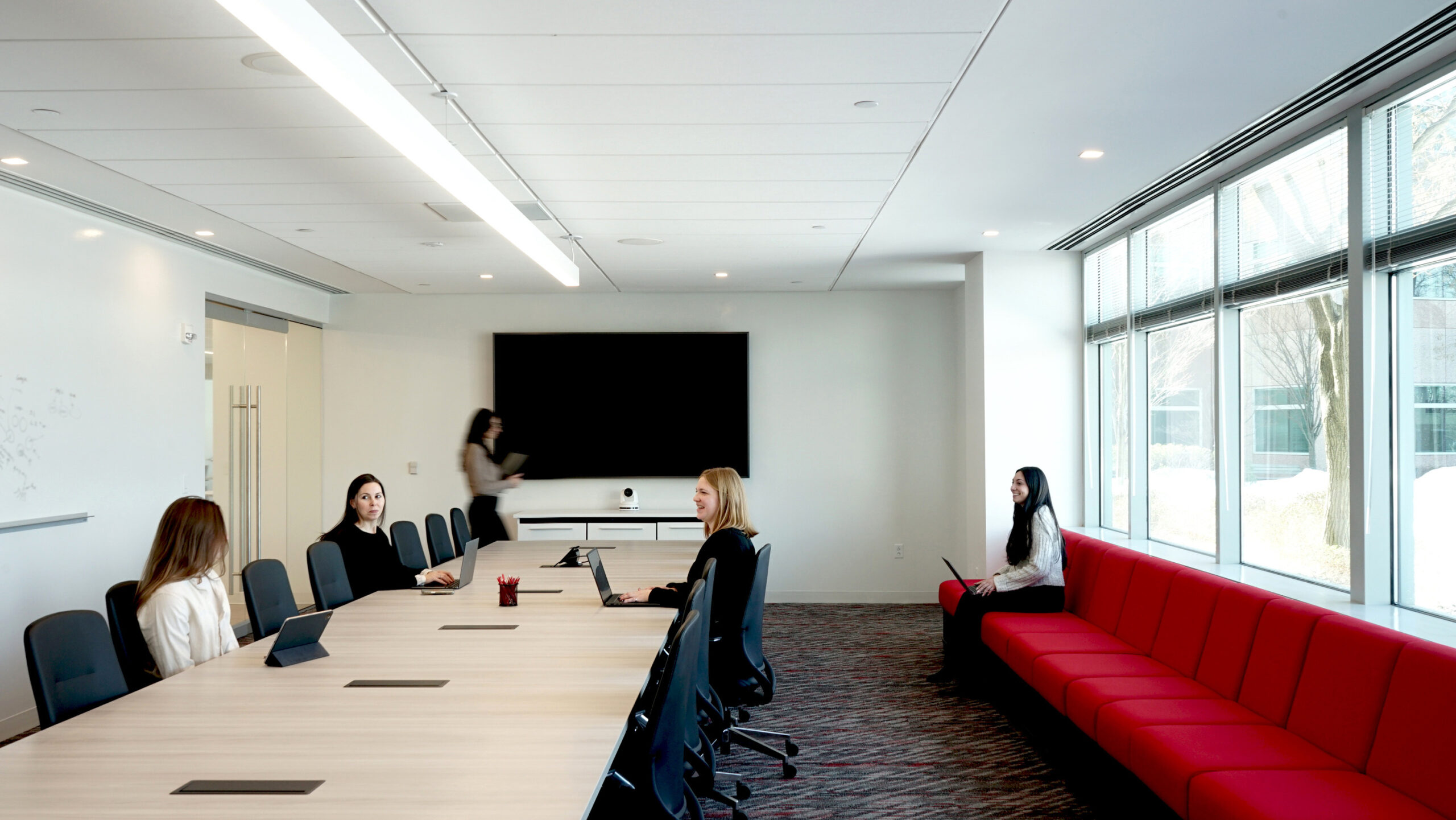 A modern conference room features a long, light wood table surrounded by ergonomic black chairs. Floor-to-ceiling windows provide natural light, enhancing the minimalist design. A vibrant red sectional sofa lines one wall, contrasting with the neutral color palette, creating a functional yet inviting space for collaboration.