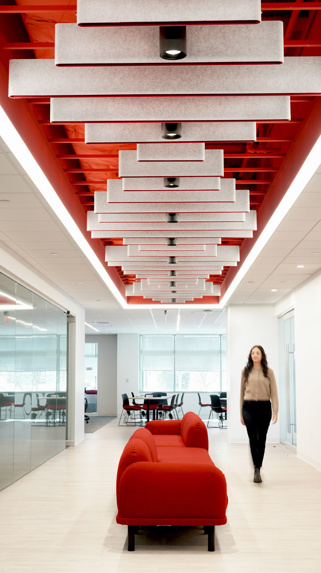 A modern office corridor showcases innovative acoustic ceiling panels in a striking red hue, complemented by sleek, white lighting. A vibrant red sofa adds a pop of color, while a glass conference room is visible in the background, enhancing the open, collaborative workspace feel.