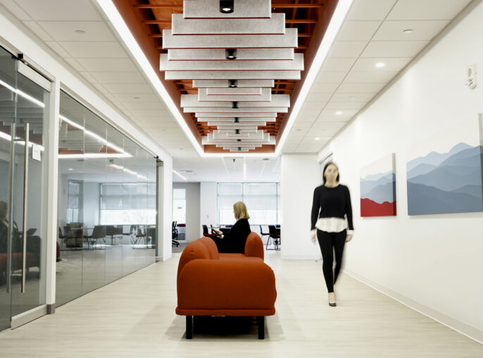 Bright, modern office hallway featuring a clean aesthetic with glass-paneled conference rooms. The striking red ceiling design contrasts against the neutral walls, while geometric acoustic panels enhance sound quality. A red sofa adds a pop of color to the seating area, complemented by abstract mountain artwork on the walls.