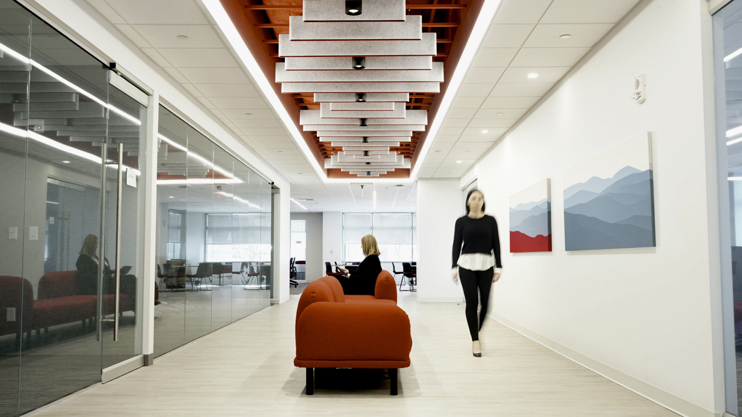 Bright, modern office hallway featuring a clean aesthetic with glass-paneled conference rooms. The striking red ceiling design contrasts against the neutral walls, while geometric acoustic panels enhance sound quality. A red sofa adds a pop of color to the seating area, complemented by abstract mountain artwork on the walls.