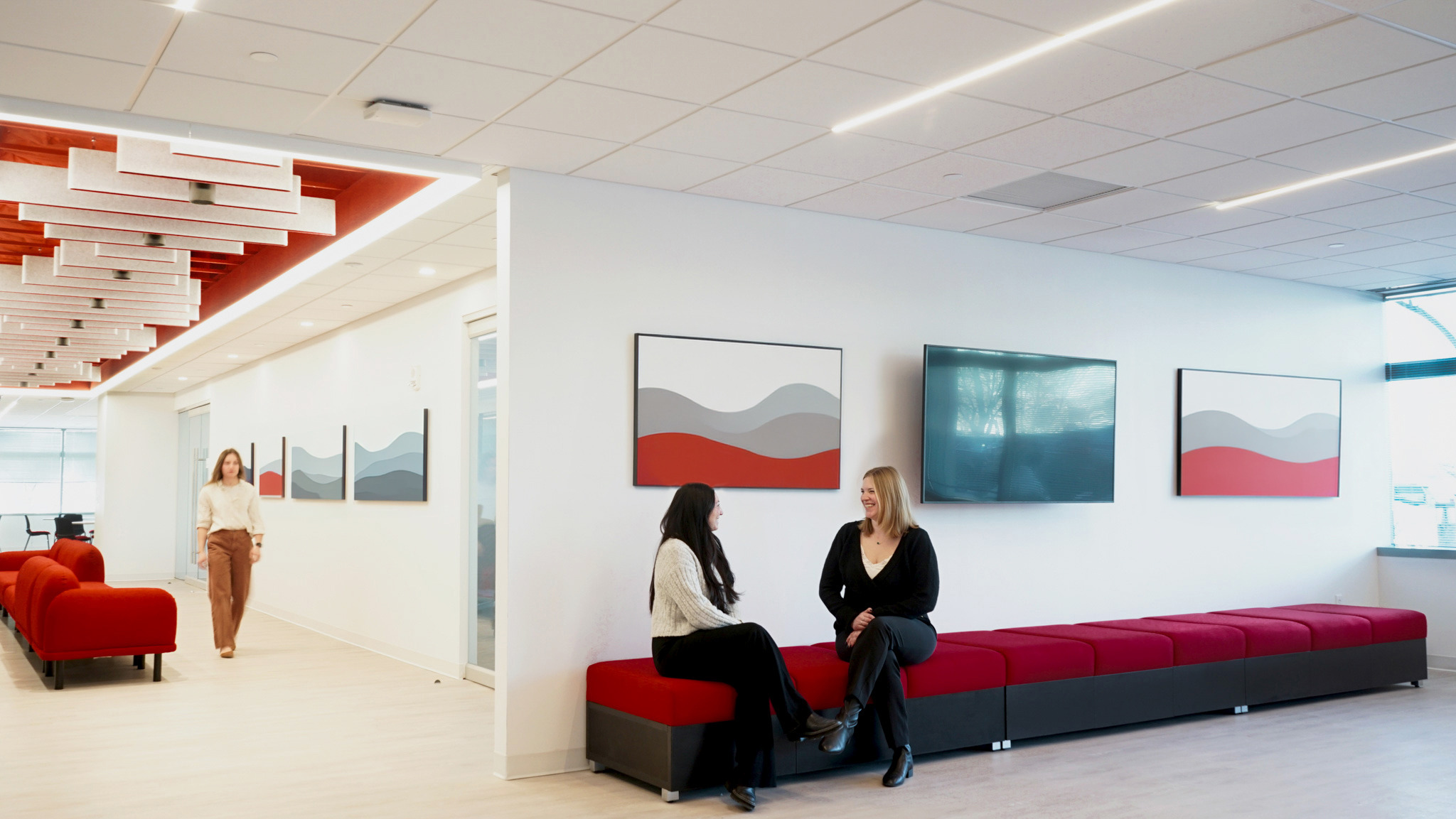 Two women engage in conversation on a striking red modular seating area, set against a clean, white wall. Abstract art pieces in subdued tones of gray and red adorn the walls, while modern lighting fixtures enhance the bright, open atmosphere of the contemporary office space. A third person walks in the background, contributing to the lively ambiance.