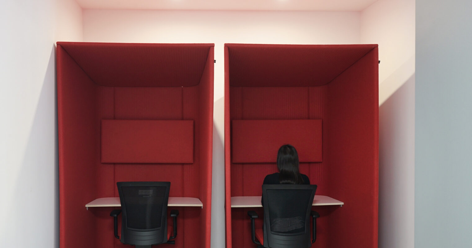 Two modern work pods featuring bold red acoustic panels provide privacy in a shared workspace. Each pod is equipped with a black ergonomic chair and a minimalistic desk, promoting focus and reducing noise. Subtle lighting enhances the contemporary aesthetic while ensuring a comfortable, productive environment.