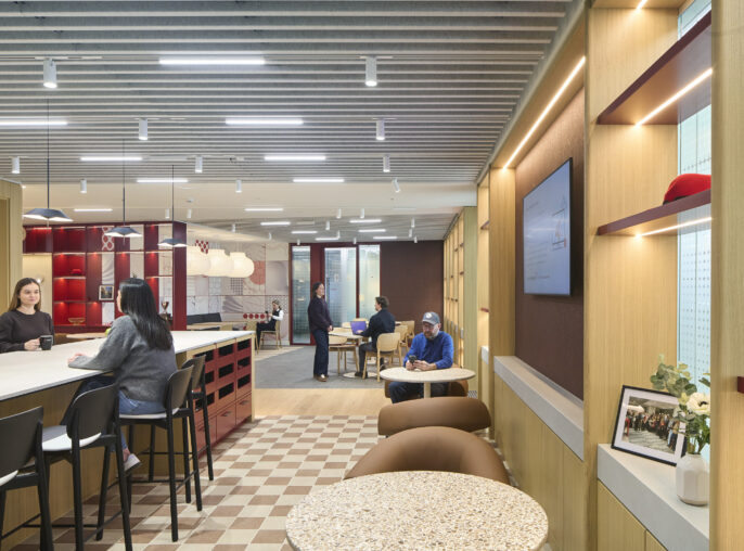 A contemporary office interior features a collaborative workspace with a light-toned wood bar and high-backed stools. Unique design elements include striking patterned wallpaper, whimsical pendant lights, and comfortably arranged seating. The layout encourages both casual interaction and focused work, enhancing a dynamic and inviting professional environment.