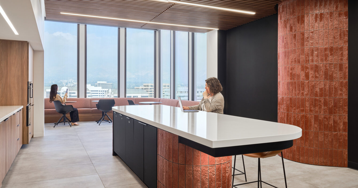 Modern office interior showcasing a sleek kitchen island with a white countertop, complemented by warm wooden accents and a striking red brick feature column. Large windows flood the space with natural light, enhancing the connection to the outdoors. Comfortable seating areas promote collaboration and relaxation.