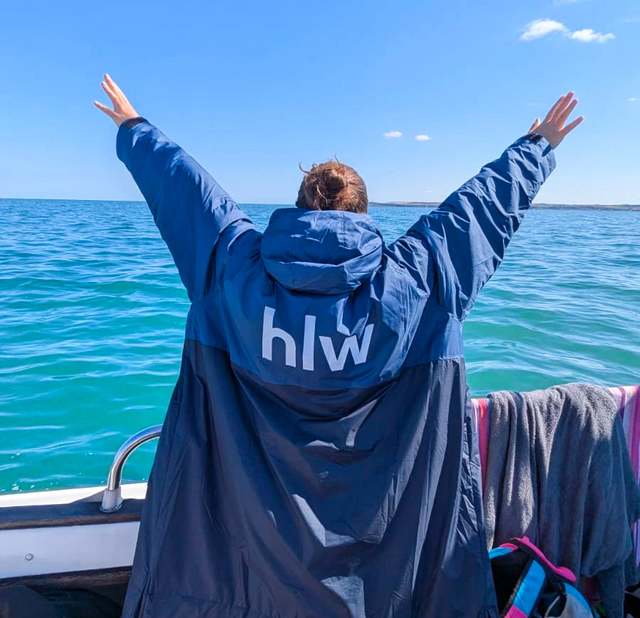 A person in a navy blue coat with the initials 'hlw' raised their arms, expressing joy against a backdrop of a clear blue sky and calm turquoise waters. The scene captures a moment of freedom, with sunlight reflecting off the water, creating a serene and lively atmosphere.