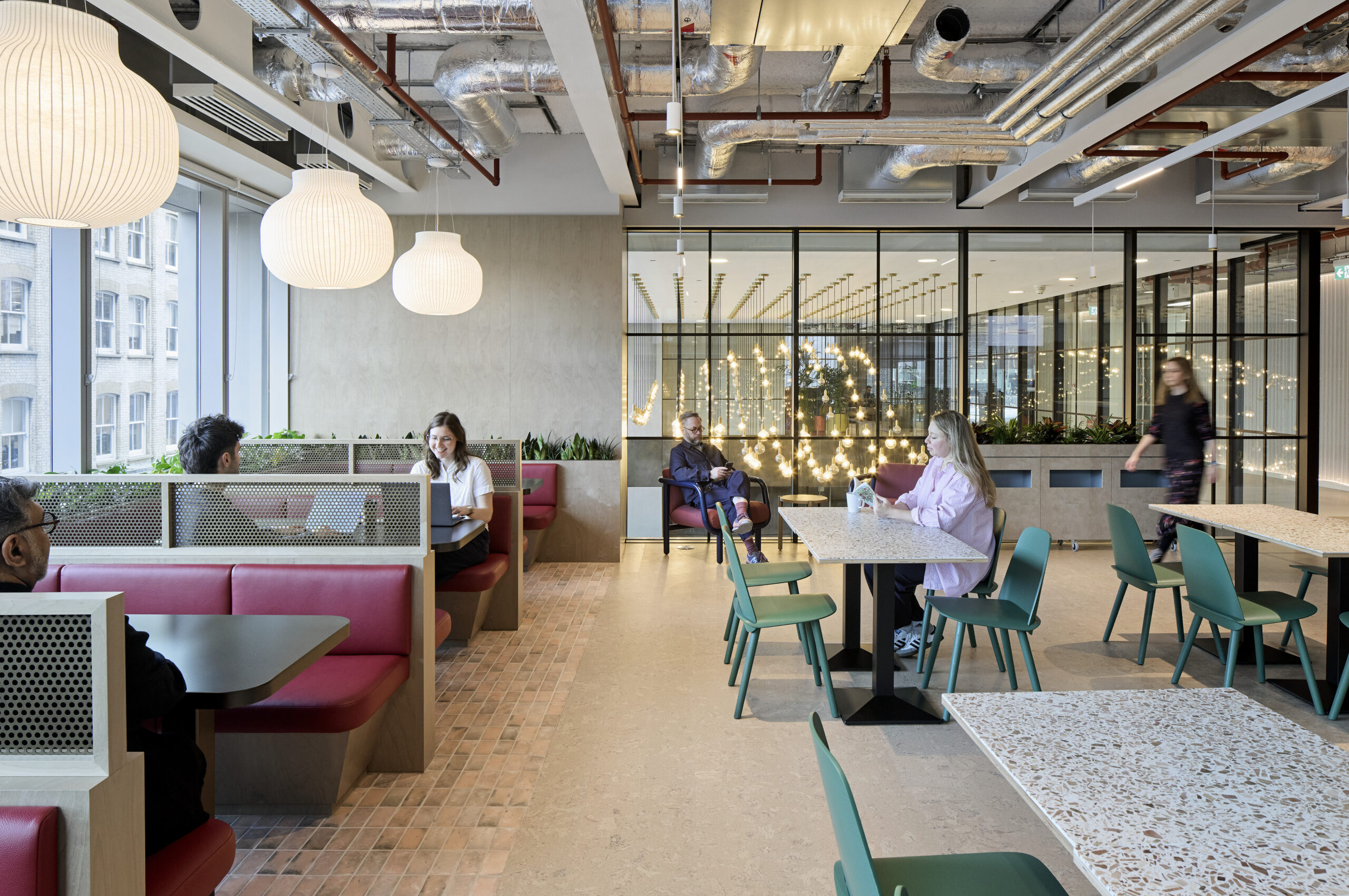 Bright and modern workspace featuring a blend of communal and private seating. Warm wood accents complement colorful booth seating and green chairs. Large windows invite natural light, while unique pendant lighting fixtures create a welcoming atmosphere. The open layout promotes collaboration and an inviting ambiance for professionals.