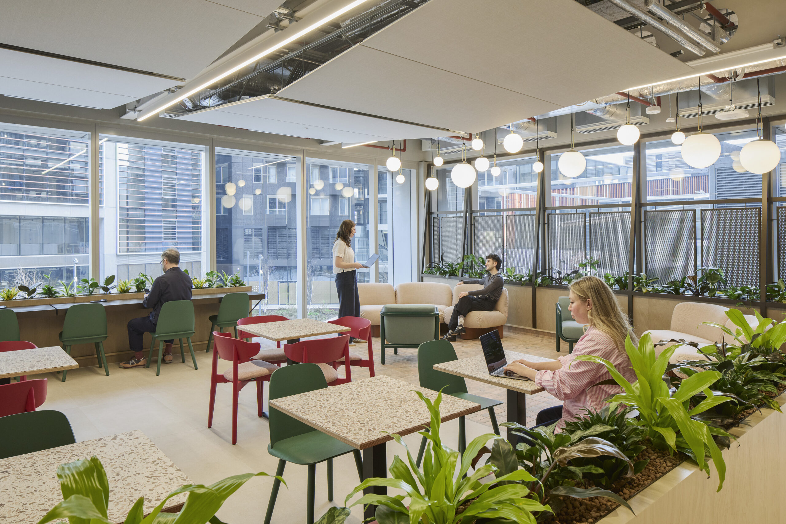 A modern co-working space featuring a blend of vibrant red and green seating arrangements. Large windows fill the area with natural light, complemented by hanging spherical light fixtures. Lush indoor plants create an inviting atmosphere, while individuals engage in collaborative work and discussions, enhancing productivity and comfort.