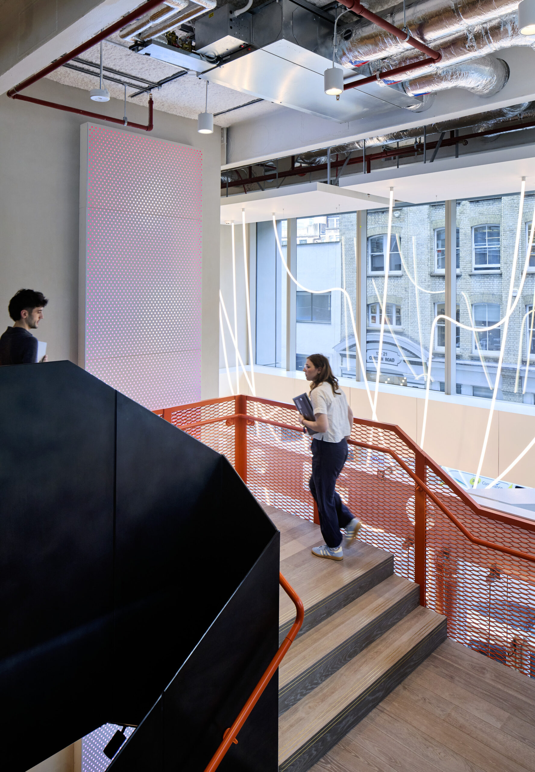 Stunning interior featuring a modern staircase with dark wood steps and a striking black handrail. A large window illuminates the space, showcasing city views and unique geometric light installations. Two professionals navigate the staircase, indicating a collaborative and dynamic work environment within the contemporary design.