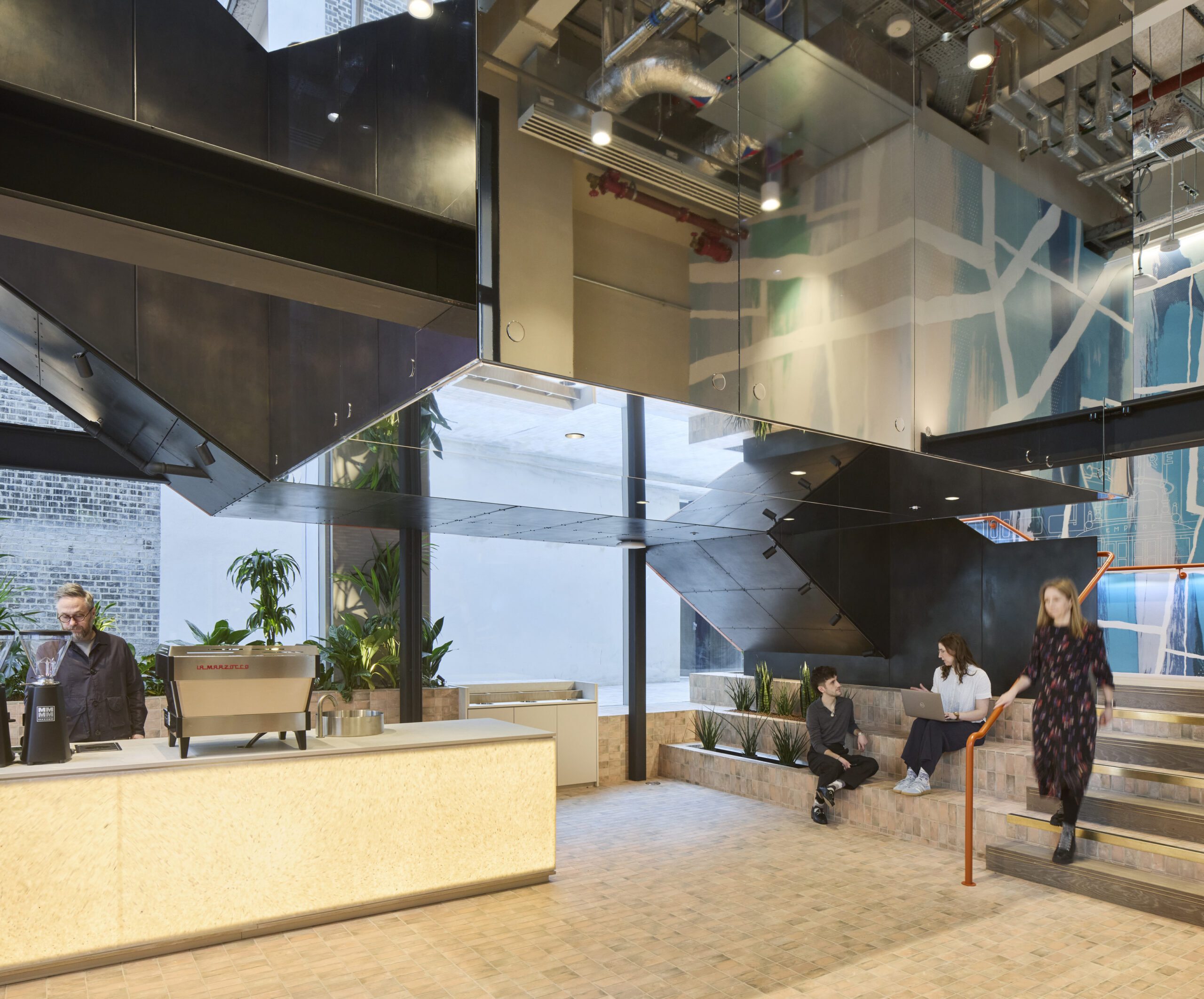 A modern commercial lobby featuring sleek black and glass elements, with an open layout and vibrant murals. A coffee bar is positioned to the left, while diverse seating areas invite collaboration. Natural light floods the space, enhancing the warm tones of the textured flooring and greenery throughout.