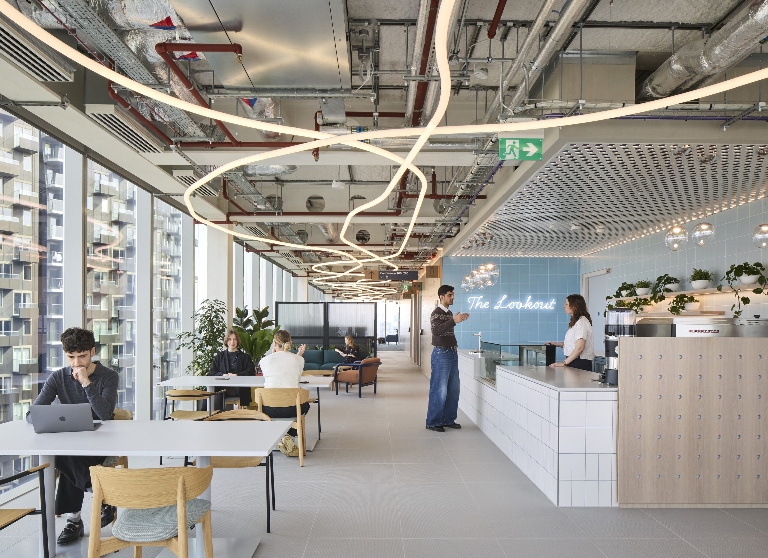 A modern open-concept office space features a sleek reception area with a blue accent wall labeled "The Lookout." Innovative light fixtures gracefully illuminate the room, while lush greenery enhances the atmosphere. Collaborative workstations and comfortable seating foster productivity and interaction among employees, highlighting contemporary workplace design principles.