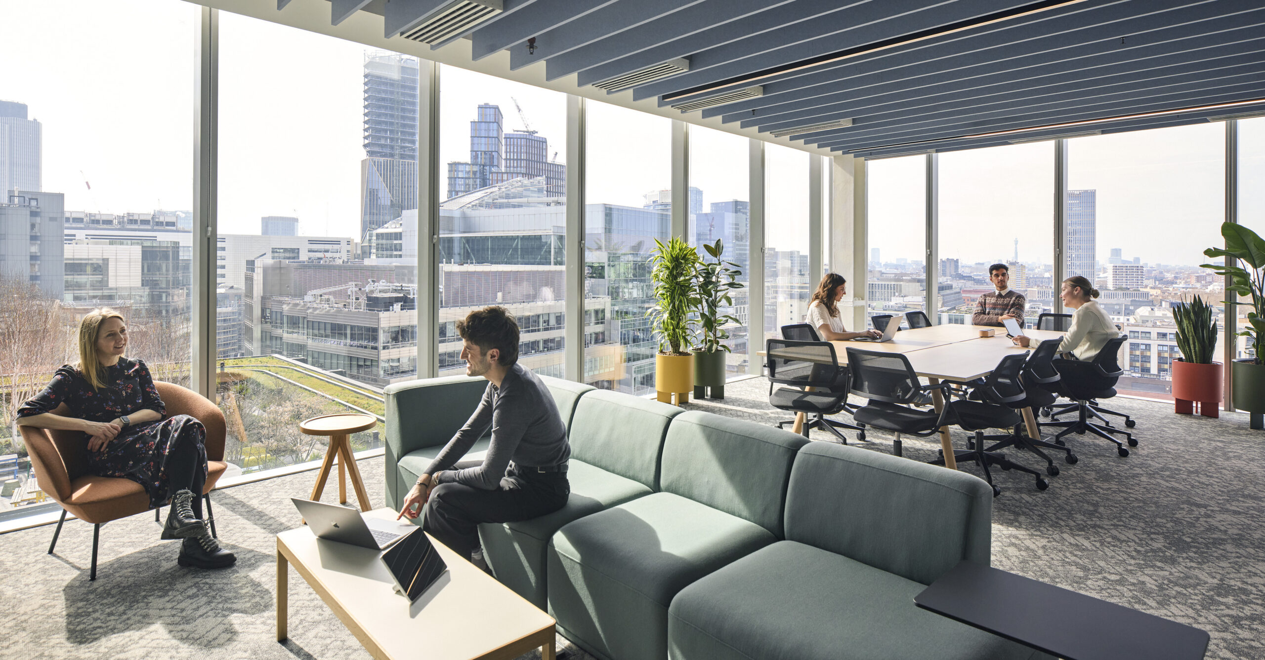Bright, modern office space featuring large floor-to-ceiling windows that offer panoramic city views. A mix of seating, including a plush sofa and minimalist chairs, fosters collaboration. Natural light enhances the open layout, with greenery adding a touch of nature, promoting a productive and inviting workplace environment.