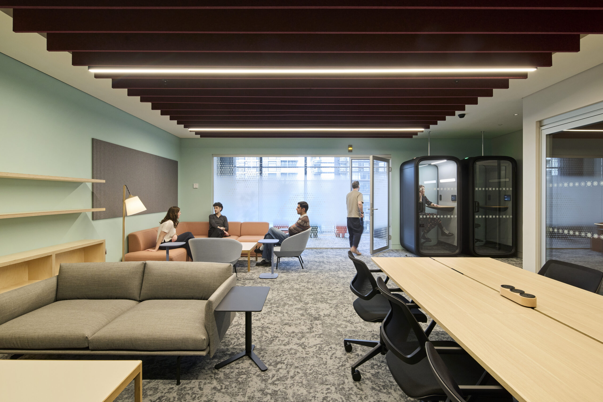 A modern office interior features a spacious, well-lit lounge area with textured walls in muted tones. Comfortable seating arrangements encourage collaboration, with soft pink and gray couches. A light wood table occupies the center, complemented by ergonomic black chairs. Glass partitions enhance openness while providing privacy for focused discussions.