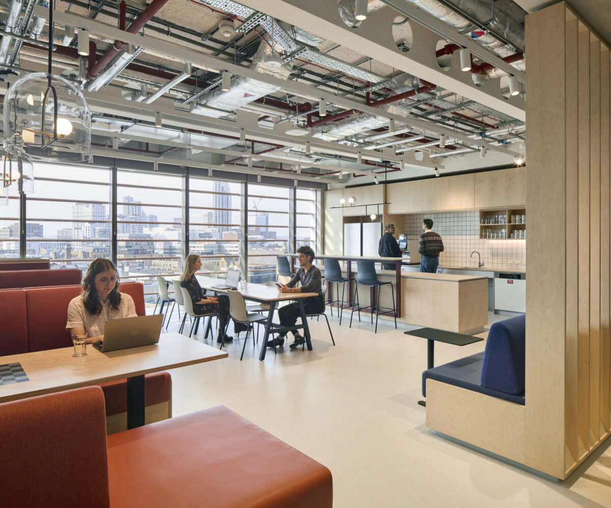 A modern office space featuring collaborative seating arrangements with vibrant orange and navy elements. Large windows illuminate the area, highlighting a contemporary kitchen backdrop and creating an open, airy atmosphere. Professionals engage in discussions, while others work on laptops, emphasizing a flexible, innovative workspace design.
