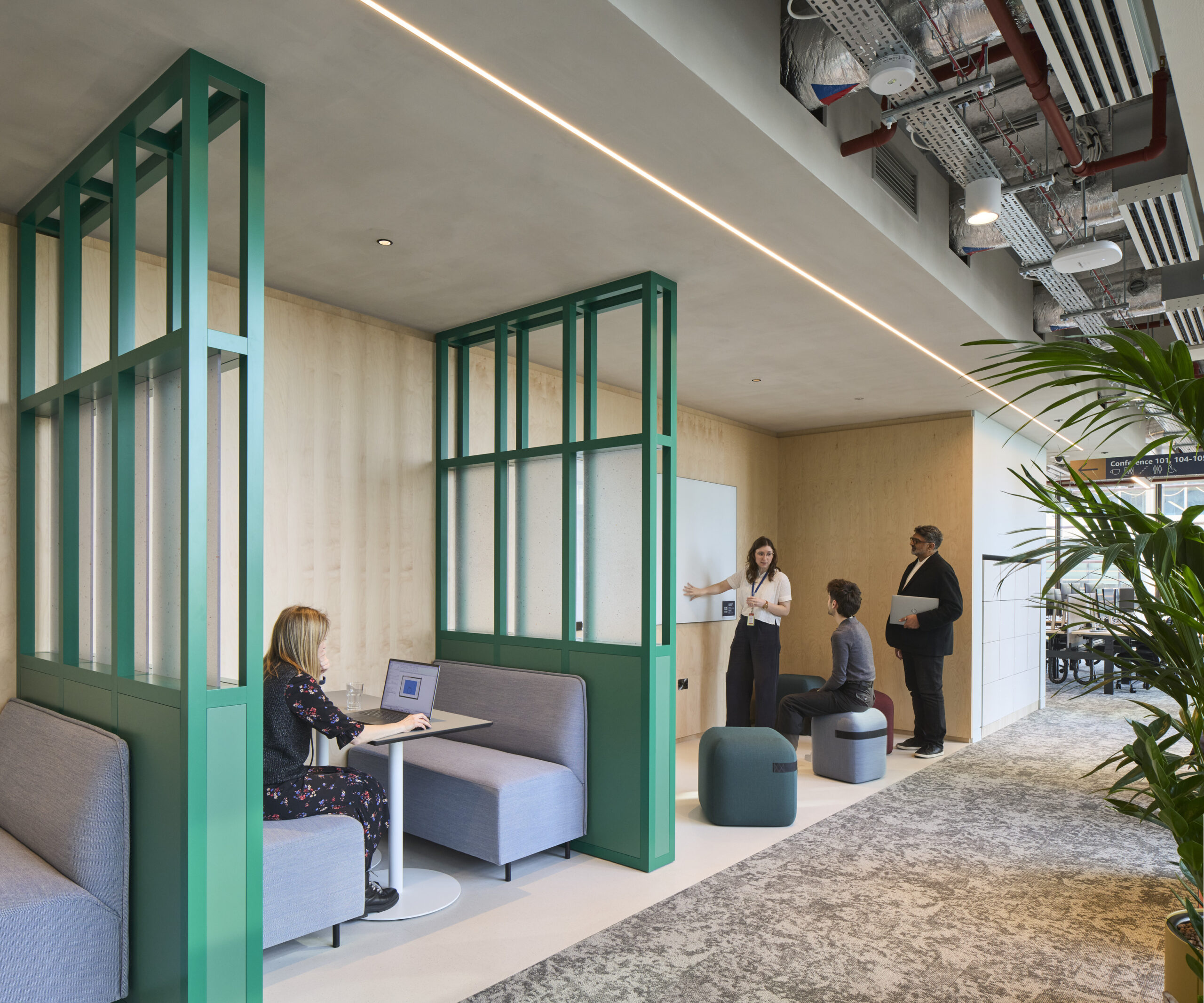 A modern workspace features acoustic partitions with vibrant green frames, creating semi-private areas for collaboration. Soft, upholstered seating invites comfort, while a light, neutral color palette enhances the airy atmosphere. Large windows allow for ample natural light, promoting a productive environment conducive to teamwork and creativity.