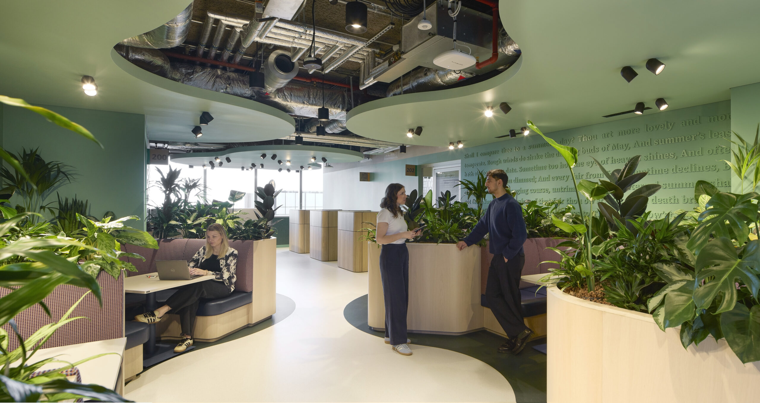A modern office interior featuring biophilic design elements. Curved green ceilings integrate with vibrant plant displays, enhancing the work environment. Informal seating areas promote collaboration, while individuals engage in work and conversation, exemplifying a sustainable, open-plan approach focused on natural aesthetics and employee well-being.