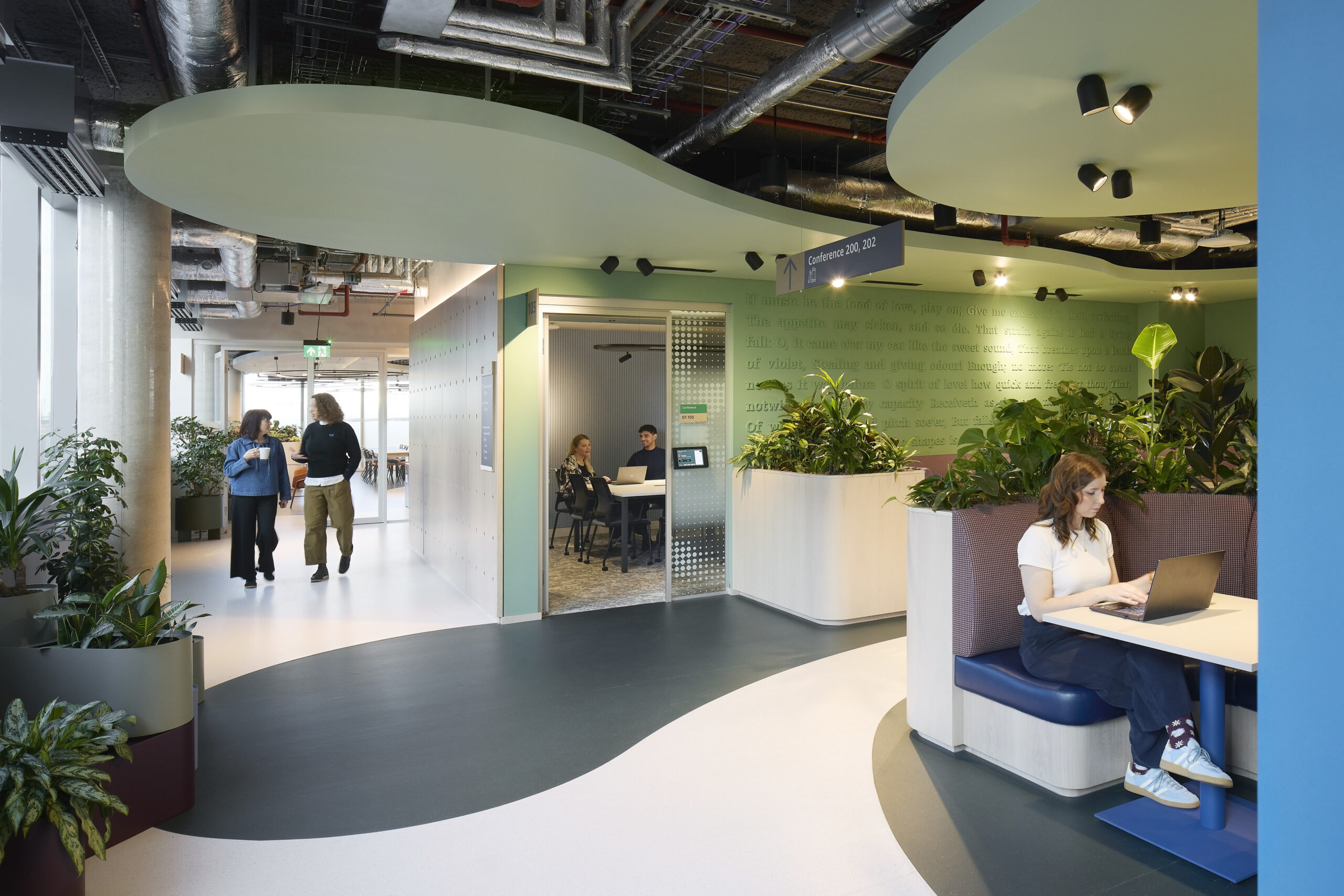 Brightly lit open office space featuring organic, wave-like green walls and a mix of indoor plants. Cozy seating areas with integrated technology invite collaboration, while glass-encased meeting rooms provide transparency. The design emphasizes biophilic elements, enhancing productivity and employee well-being through nature-inspired aesthetics.