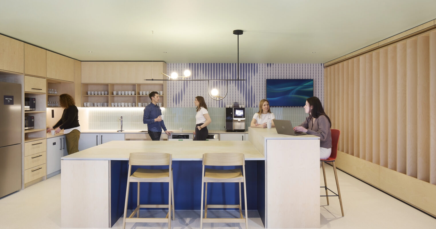 A modern kitchen designed for collaboration features sleek cabinetry, a spacious island with blue accents, and minimalist bar stools. Natural light floods the space through large windows, enhancing the warm wood tones and contemporary decor. Group interactions promote a sense of community in this functional, stylish environment.