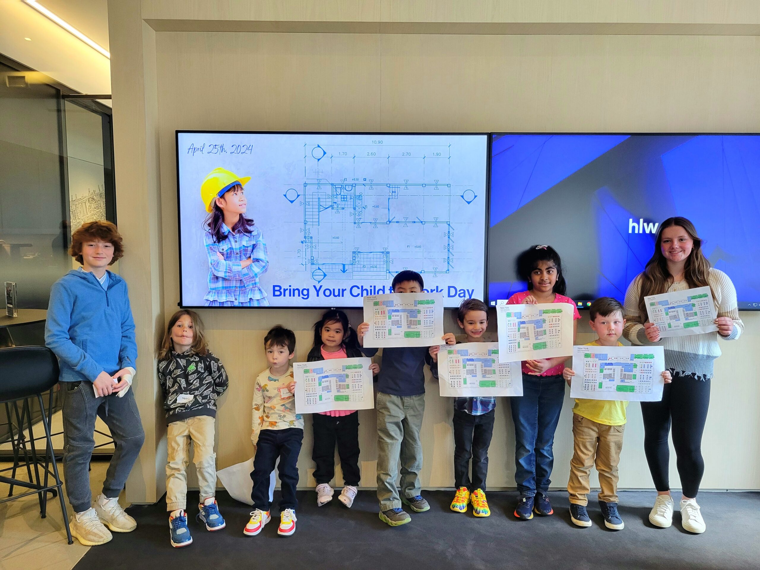 Children display completed floor plan activities during Bring Your Child to Work Day in a modern office environment, standing in front of digital screens featuring architectural drawings and workplace design concepts.