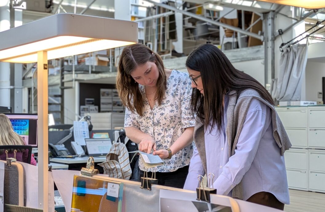 Two team members are reviewing interior design materials and project documents at a workstation within a modern open-plan studio, illustrating mentorship, knowledge sharing, and professional development in a collaborative workplace.