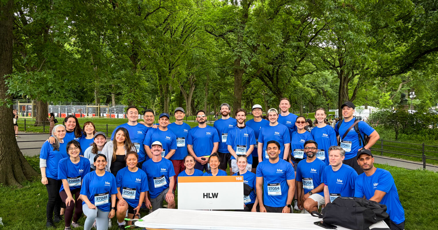 HLW team members wearing blue shirts gather in a tree-lined urban park during a JP Morgan race, highlighting employee engagement, wellness, and workplace culture.