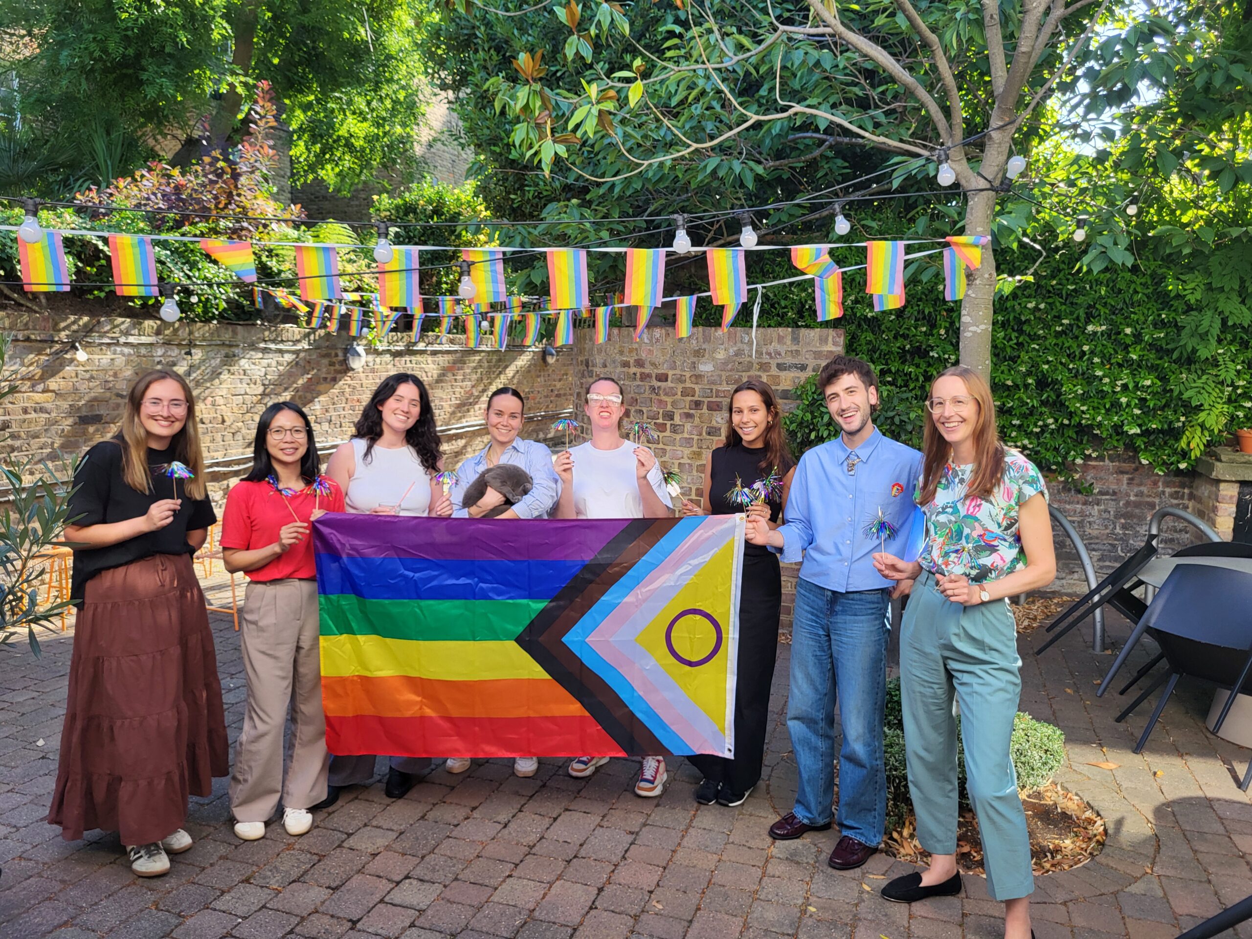 HLW team members gather in an outdoor courtyard decorated with Pride flags, holding a Pride flag and celebrating inclusion, community, and workplace culture.