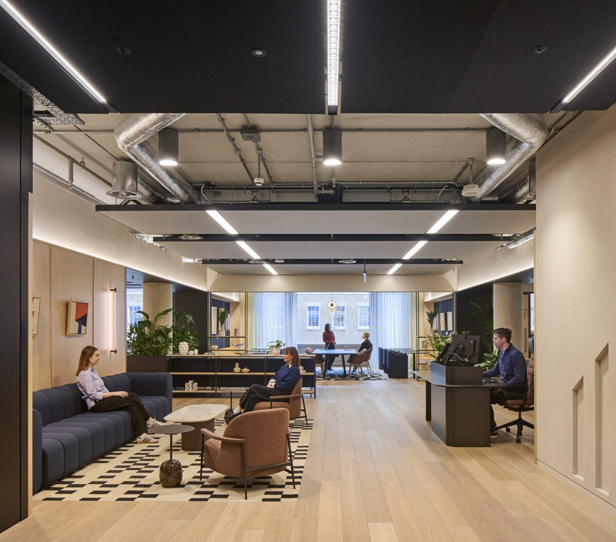 Open-plan office lounge with exposed ductwork, dark ceiling panels, light oak flooring, navy sofas, a geometric rug, plants, and employees seated in a layered reception and collaboration area.