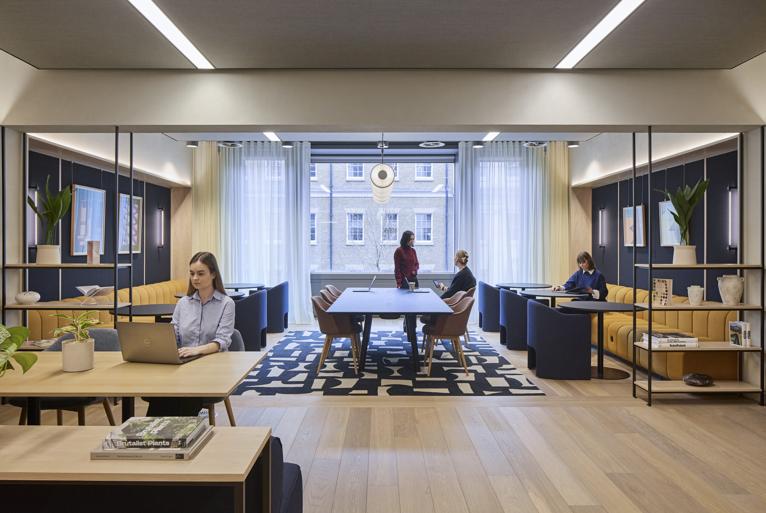 Collaborative office lounge with a central wood meeting table, mustard banquettes, navy lounge chairs, open shelving, framed artwork, plants, and employees working beside a large window.