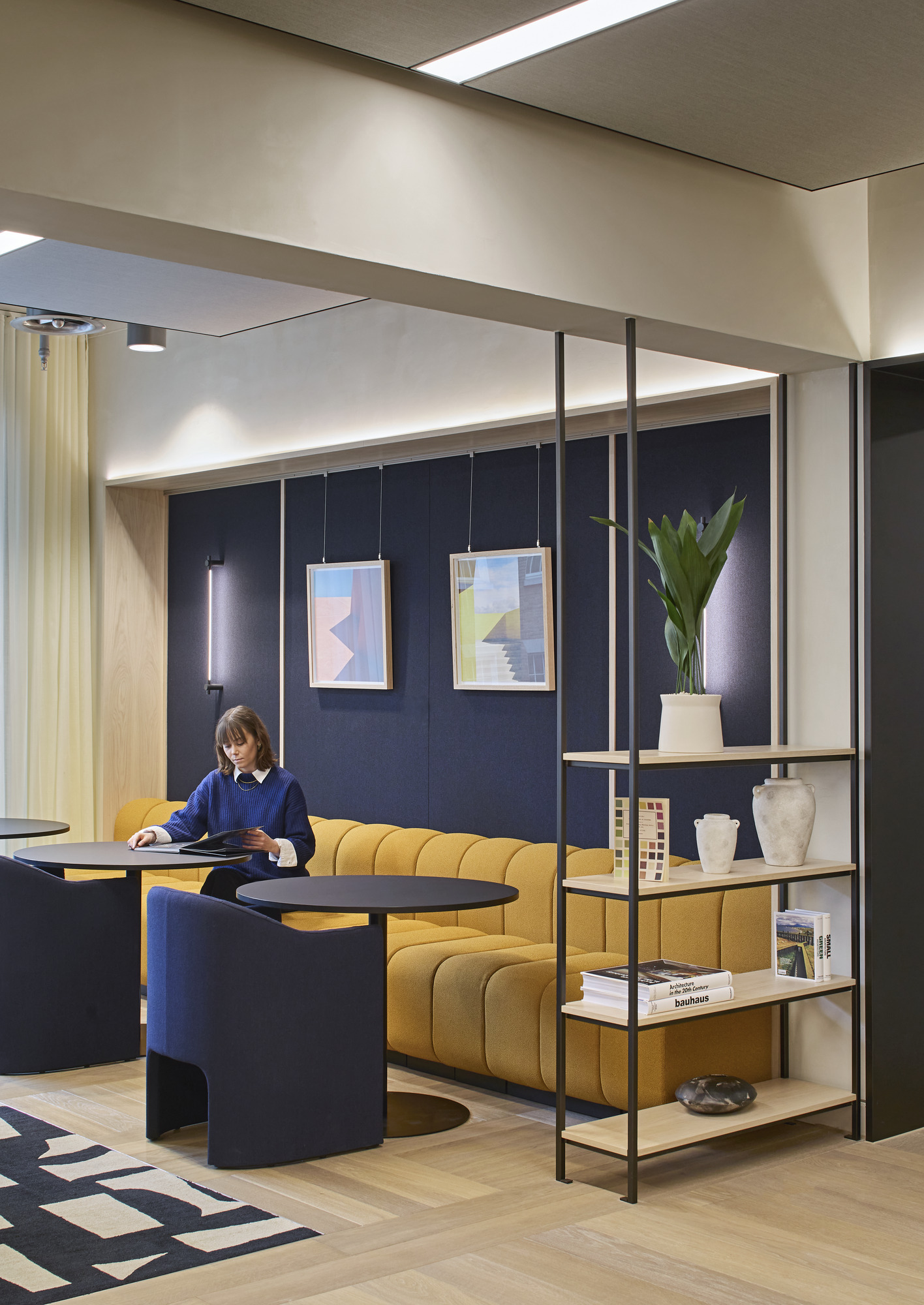 Office lounge with a mustard upholstered banquette, navy armchairs, dark acoustic wall panels, framed geometric artwork, open wood shelving, plants, and integrated linear lighting.