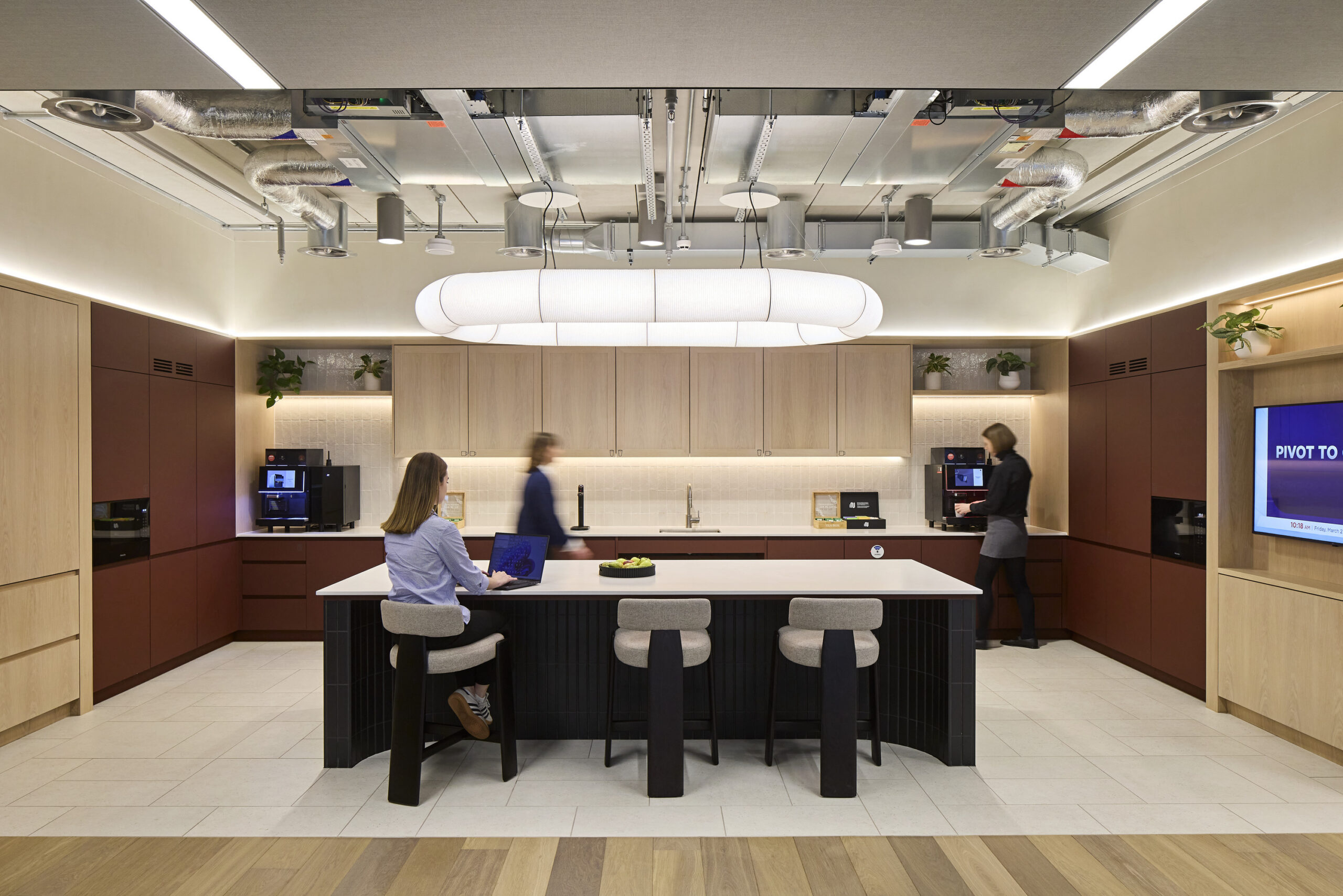 Modern office kitchen with oak wall cabinets, dark red base cabinetry, a large island with a white countertop, integrated appliances, exposed mechanical services, and pendant lighting.