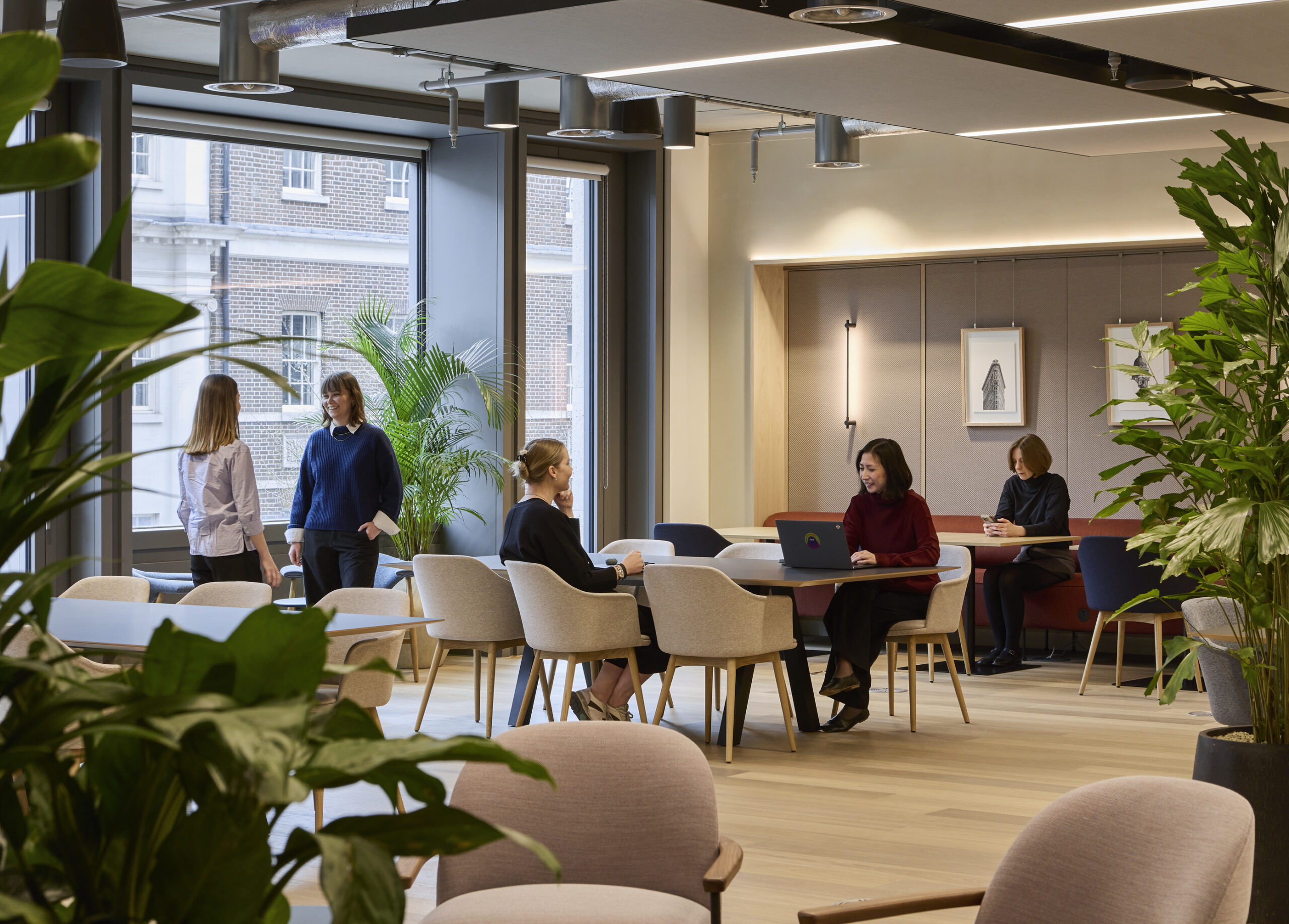 Office collaboration area with large windows, light oak flooring, pale wood tables, upholstered chairs, planted interiors, and employees working and conversing around shared dining tables.