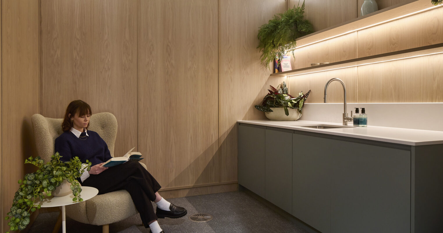 Quiet office refreshment room with full-height oak cabinetry, a gray kitchenette with sink, illuminated display shelves, potted plants, and a woman reading in a lounge chair.
