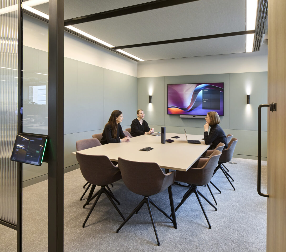 Glass-enclosed conference room with a large wood meeting table, upholstered chairs, a wall-mounted display, video-conferencing equipment, and three people seated in discussion.
