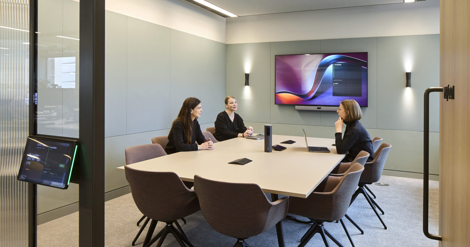 Glass-enclosed conference room with a large wood meeting table, upholstered chairs, a wall-mounted display, video-conferencing equipment, and three people seated in discussion.