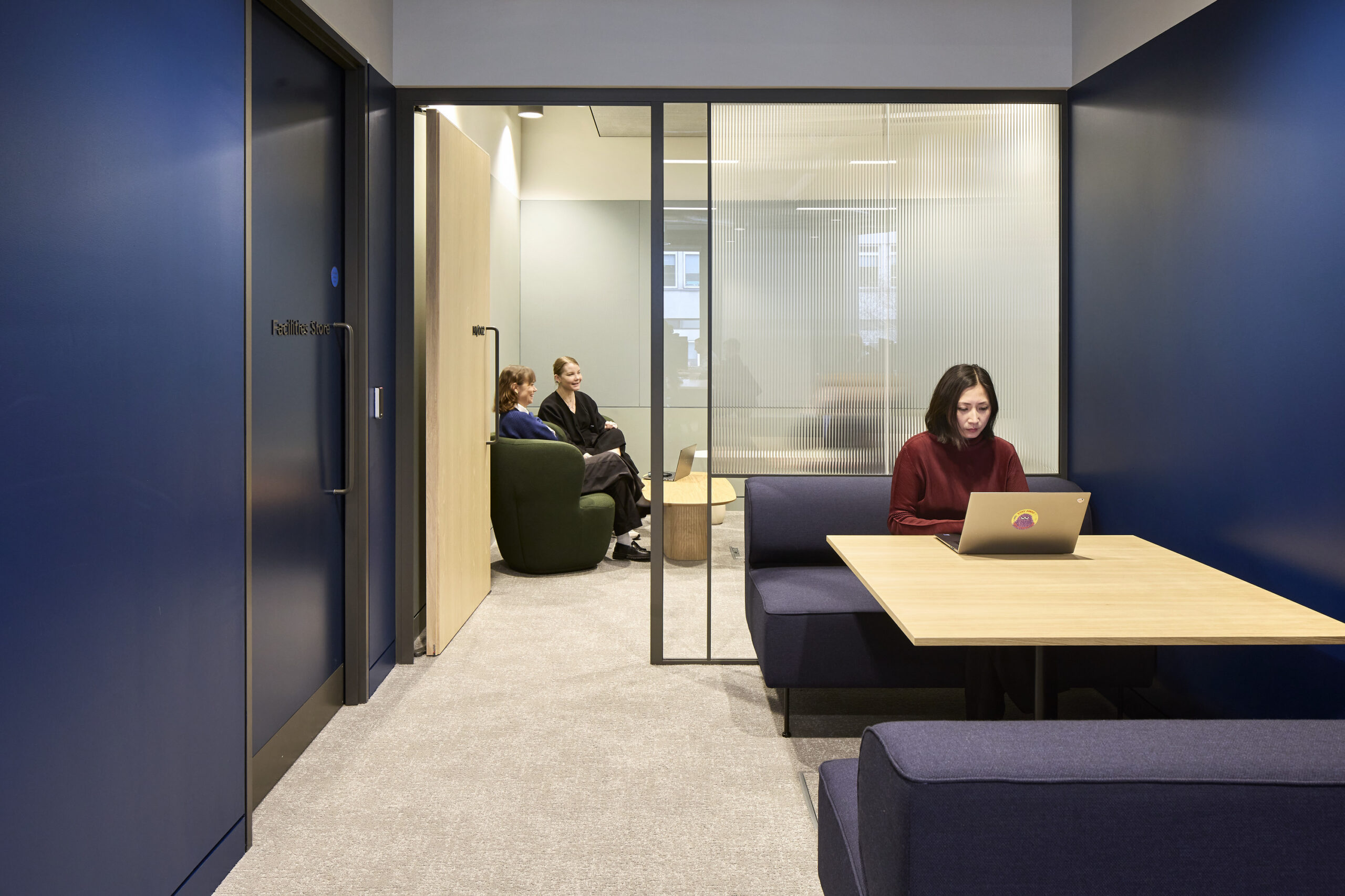 Office breakout area with deep blue walls, a woman working at a laptop in the foreground, and two people seated in a lounge space behind fluted glass partitions.