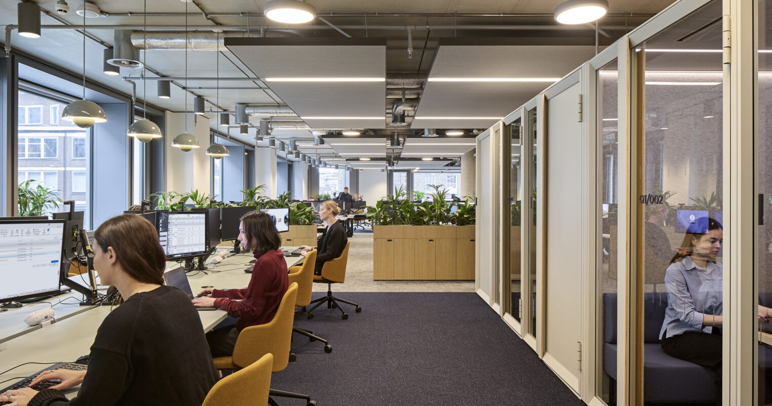 Open-plan office with exposed mechanical services, suspended acoustic ceiling panels, linear lighting, rows of workstations, mustard task chairs, planted storage units, and glazed meeting rooms.