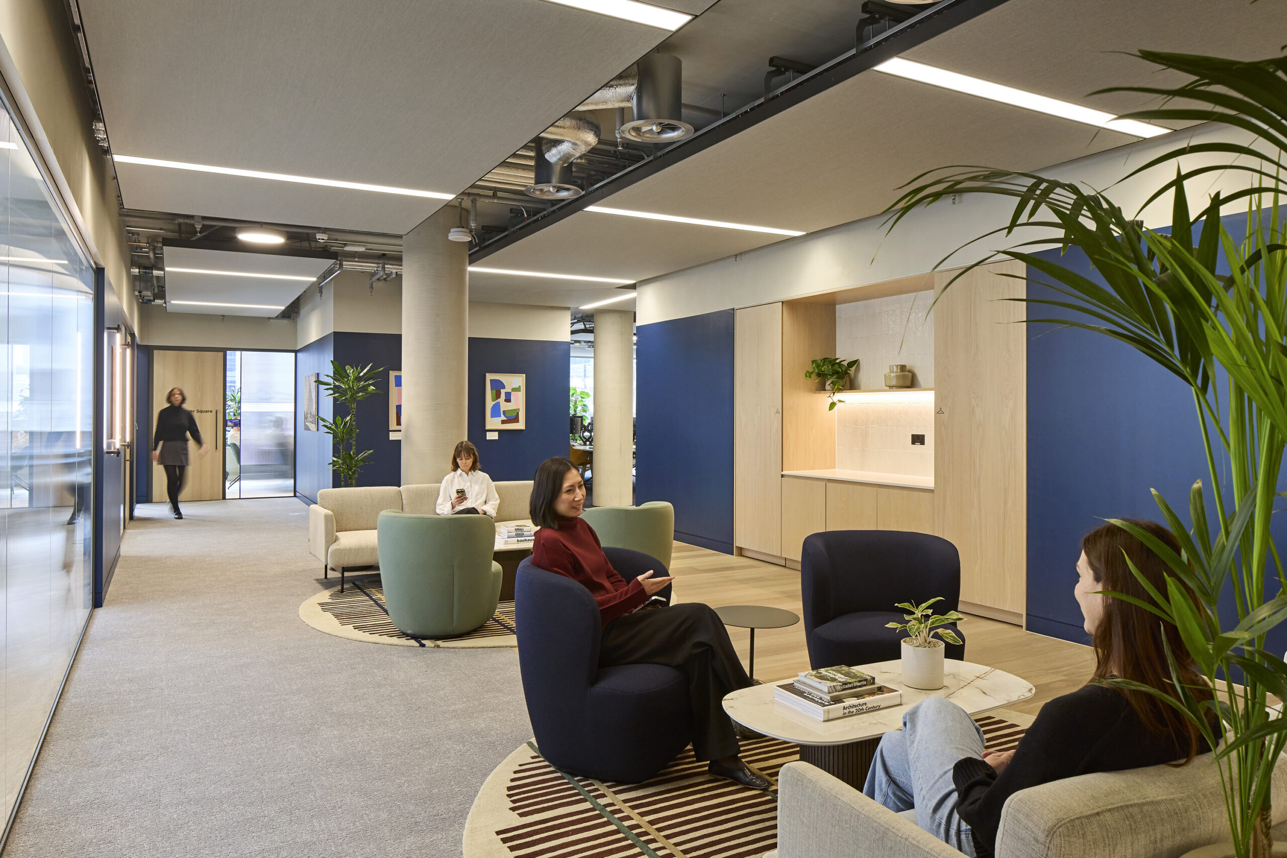 Office lounge and corridor with blue wall panels, light oak cabinetry, exposed services, acoustic ceiling panels, patterned rugs, upholstered lounge chairs, plants, and employees conversing.