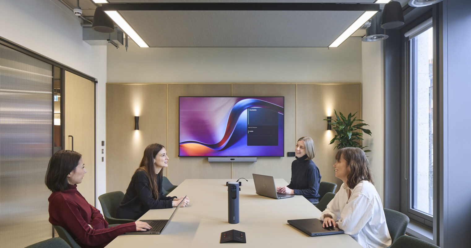 Conference room with a large pale wood table, green upholstered chairs, a wall-mounted display, wood-paneled feature wall, integrated sconces, and windows along one side.
