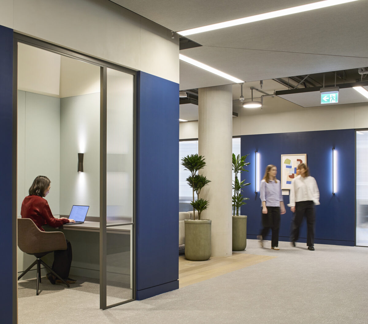 Office corridor with a glass-enclosed workspace, blue wall panels, concrete columns, potted plants, carpet flooring, suspended acoustic ceiling panels, and employees walking through the space.