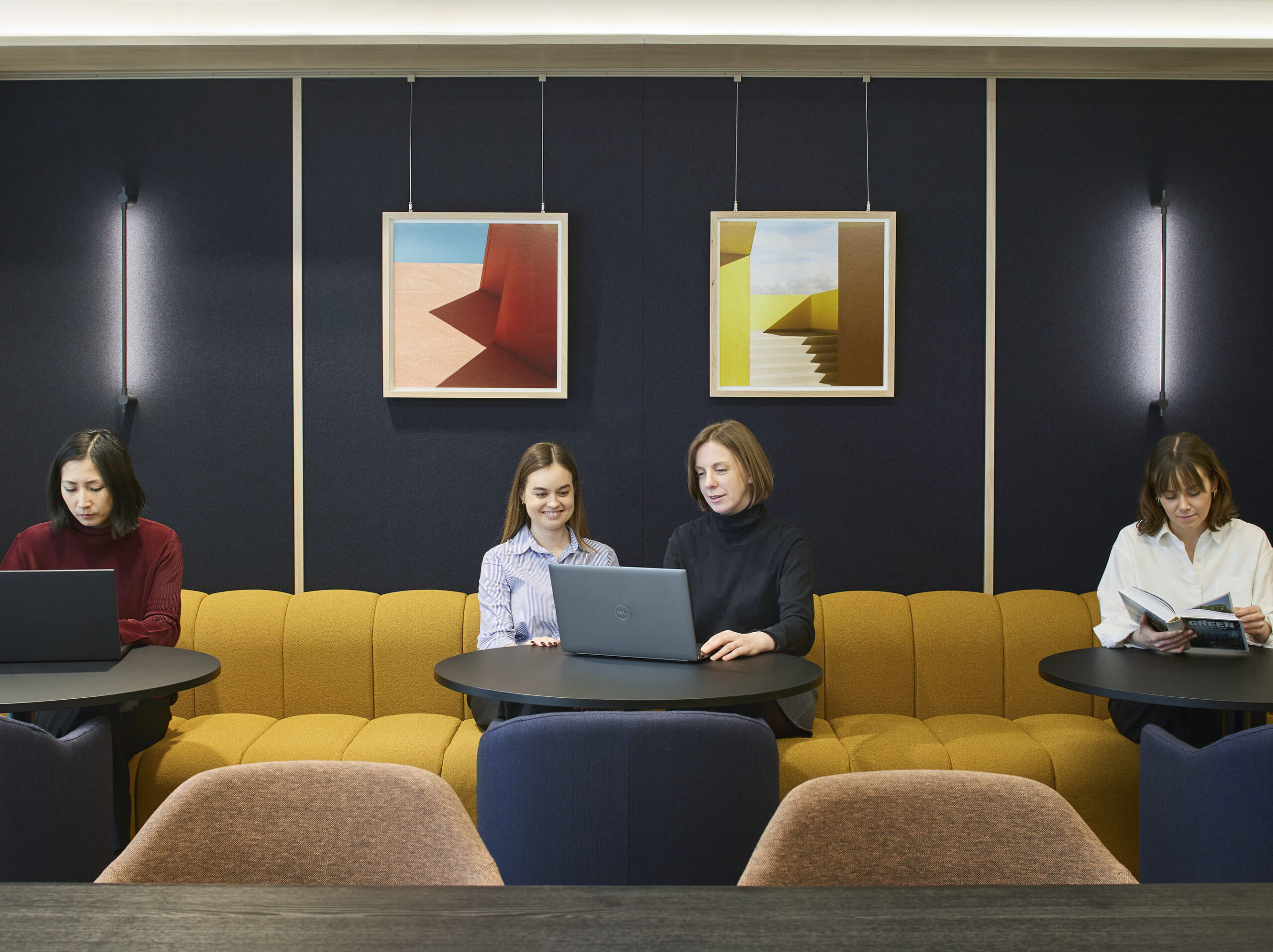 Collaborative lounge with a mustard upholstered banquette, dark acoustic wall panels, geometric artwork, black round tables, navy lounge chairs, and four people working or reading.